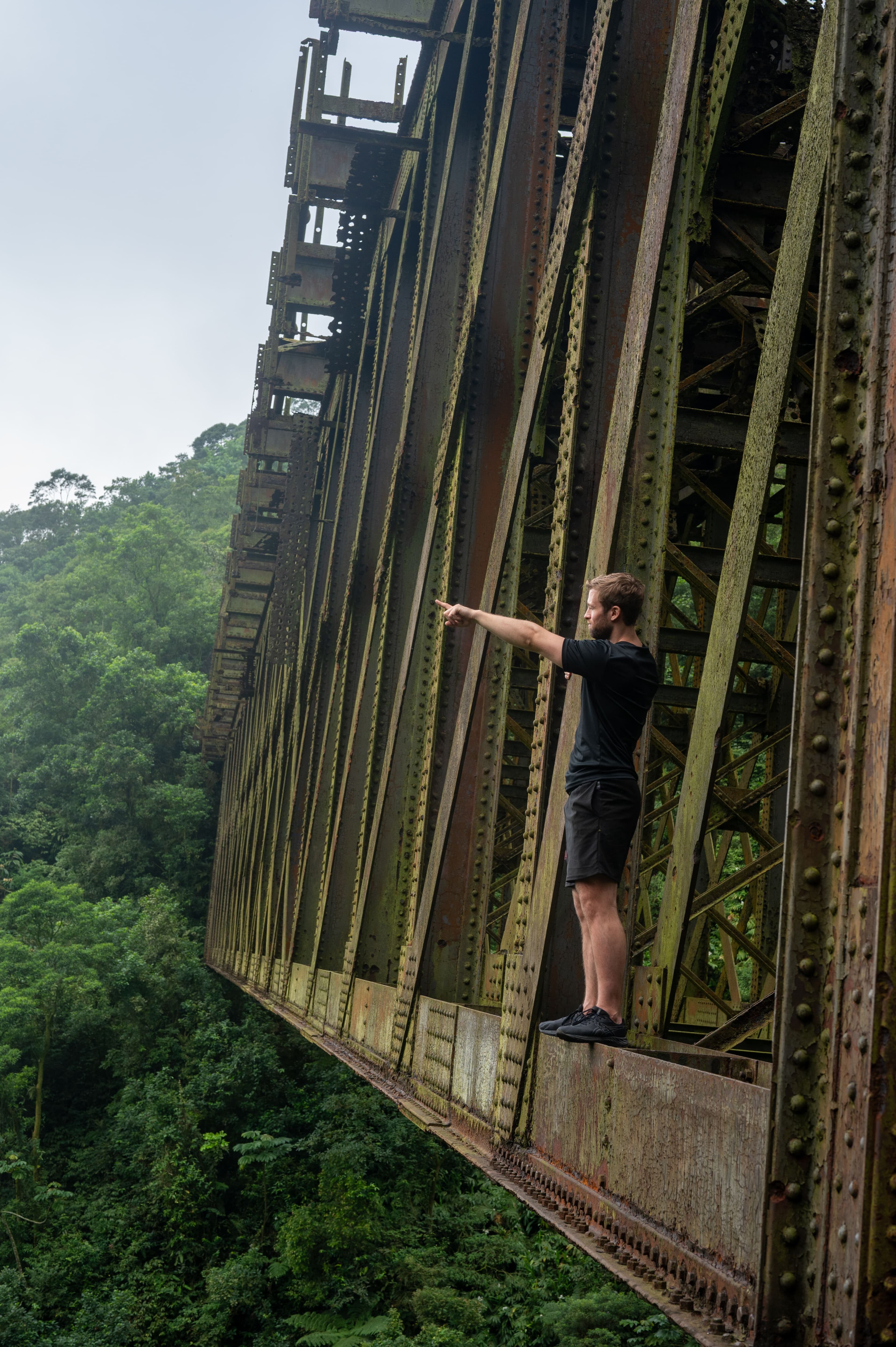 Man in black points from a massive, rusty bridge overlooking a dense green forest.
