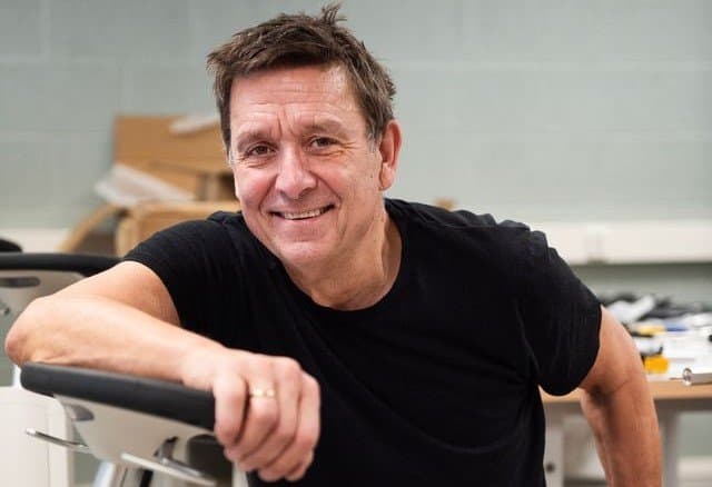 Smiling middle-aged man in a black t-shirt leaning on the handlebars of exercise equipment.