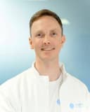 Smiling male doctor with short brown hair wearing a white lab coat against blue background.
